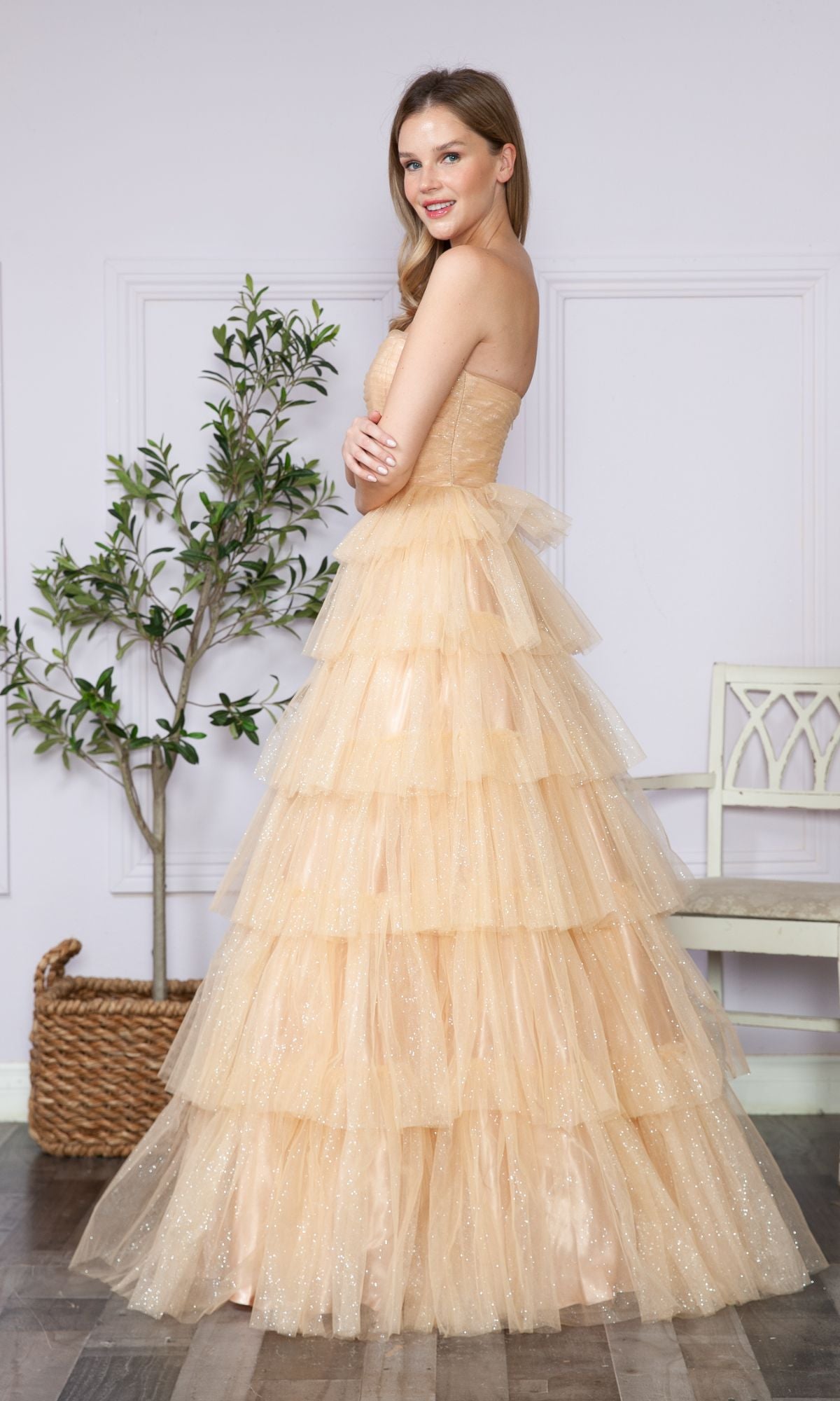 A woman stands indoors beside a potted plant and white chair, smiling in the Strapless Long Tiered Glitter Prom Ball Gown 9386—an elegant peach-colored, strapless dress with layered tulle.